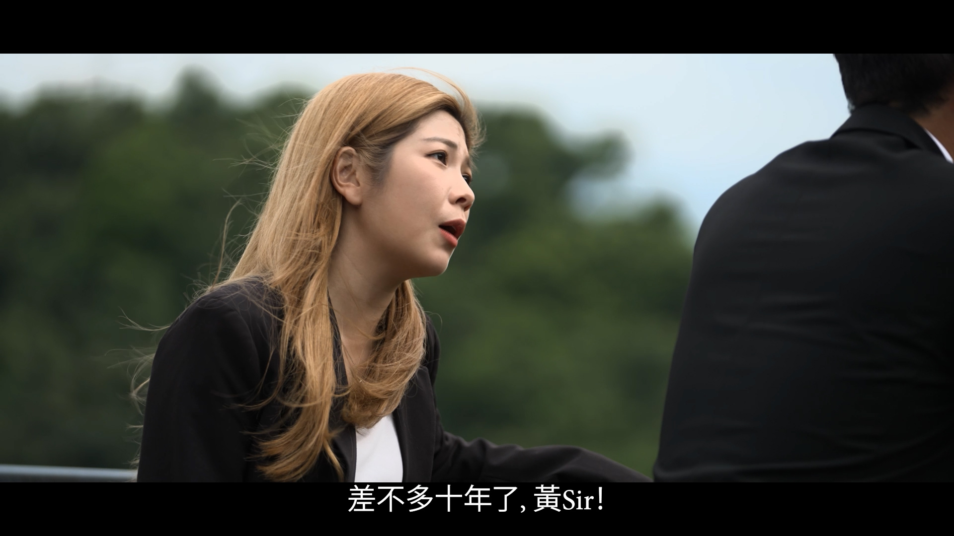 A woman with long blonde hair, wearing a black blazer, speaks outdoors with trees in the background. A man in a suit sits beside her. Chinese and English subtitles appear at the bottom of the image.