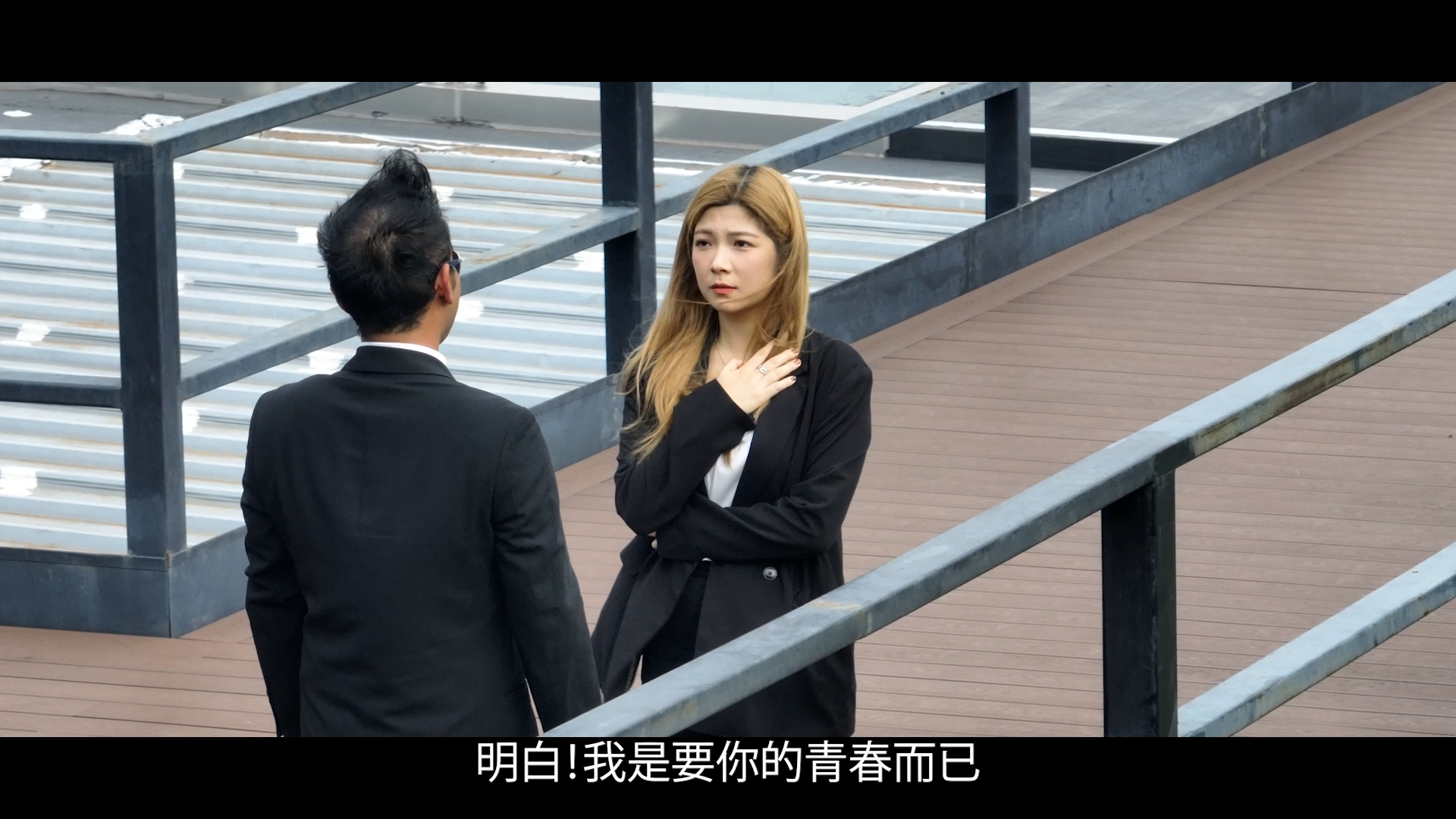A man and a woman stand facing each other outside on a wooden deck. The woman has one hand on her chest and looks concerned, while the man stands with his back to the camera. Chinese subtitles appear at the bottom.