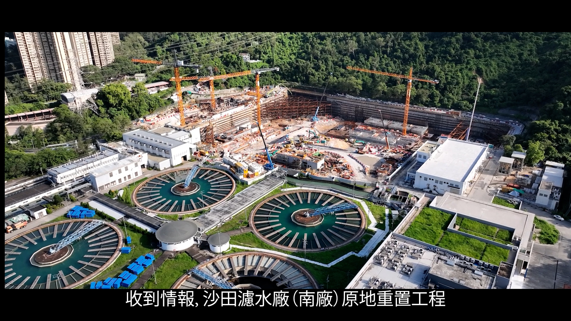 Aerial view of a large water treatment facility under construction, with cranes, circular water tanks, and nearby buildings surrounded by greenery. Chinese text appears at the bottom of the image.