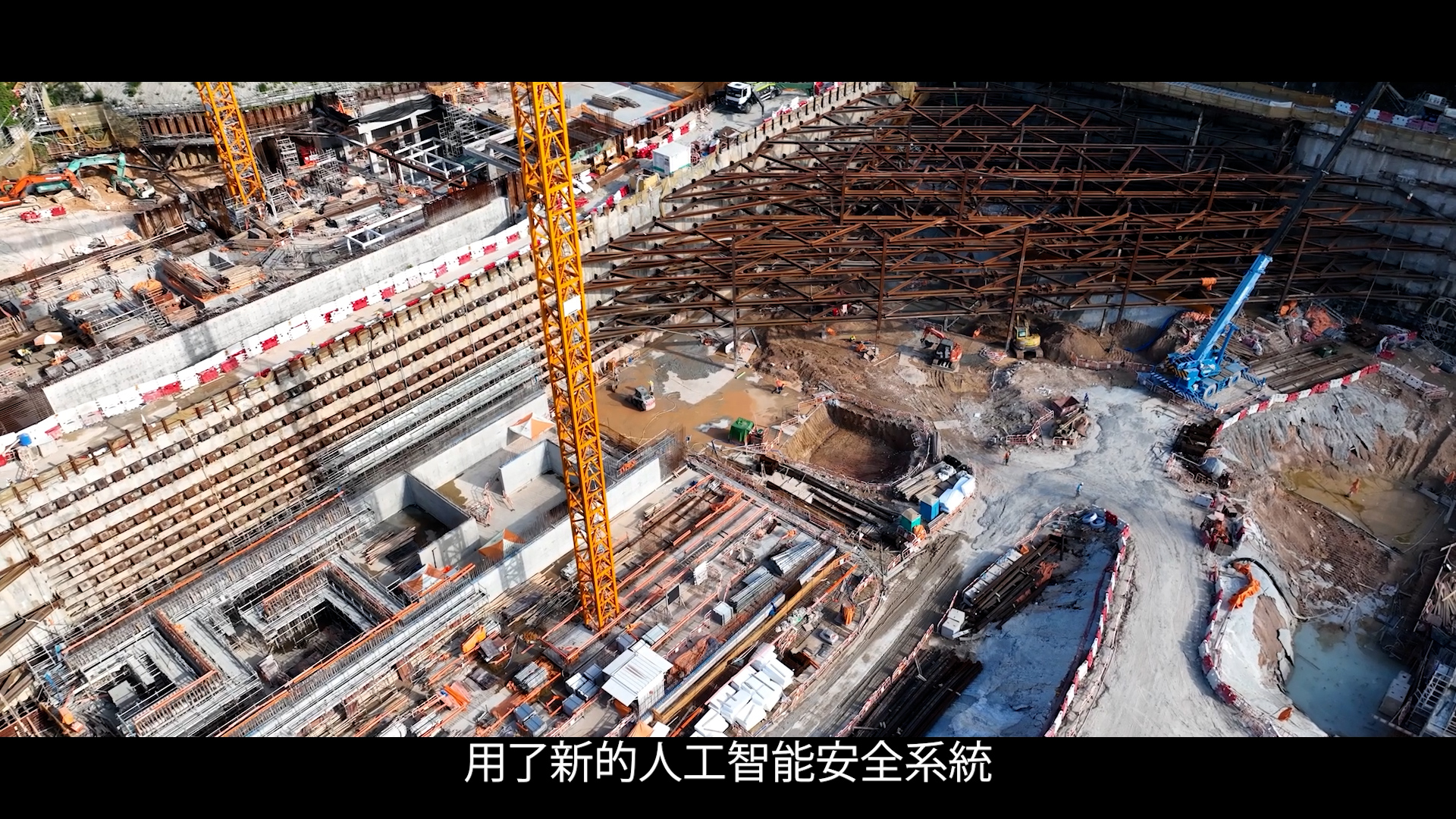 A large construction site with cranes, steel frameworks, and equipment. Workers and building materials are visible. Chinese subtitles at the bottom read: "用了新的人工智能安全系统" ("A new AI safety system is used").