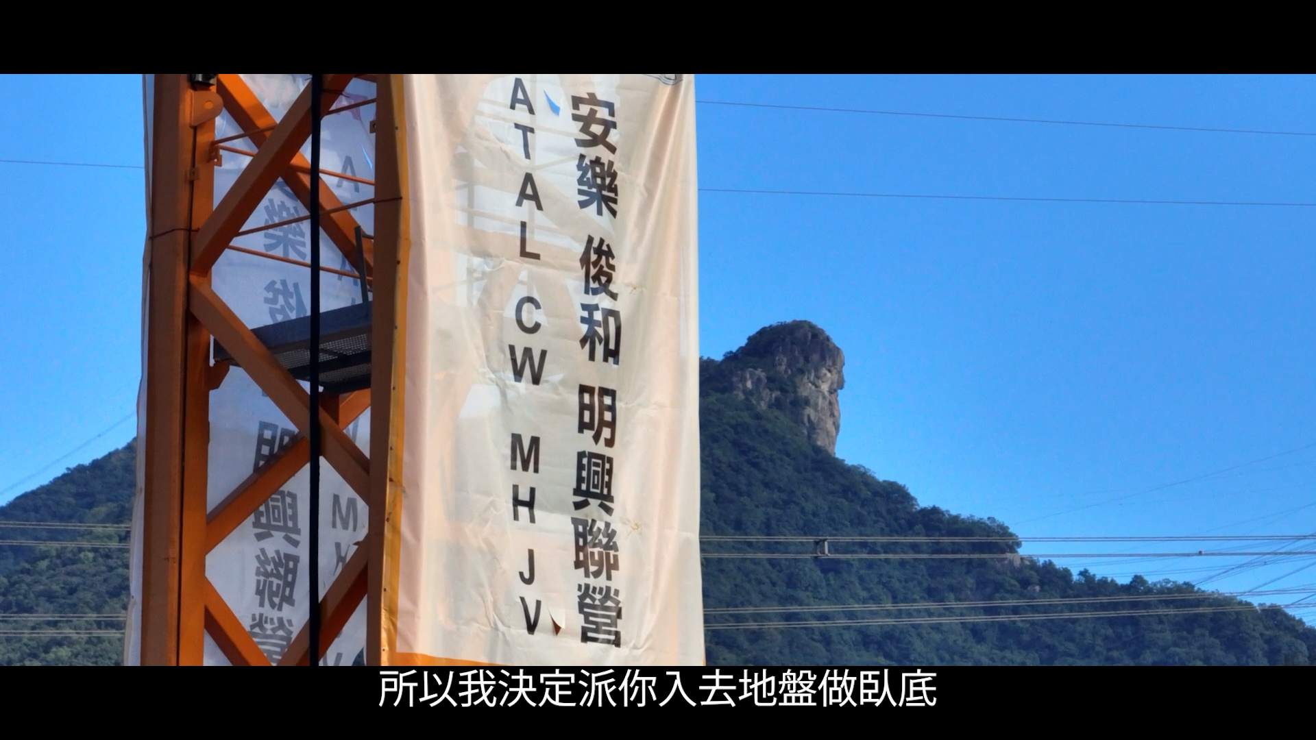 A vertical banner with Chinese and English text hangs on a metal structure in the foreground. In the background, there is a mountain with a distinct rocky peak under a clear blue sky. Chinese subtitles appear at the bottom.