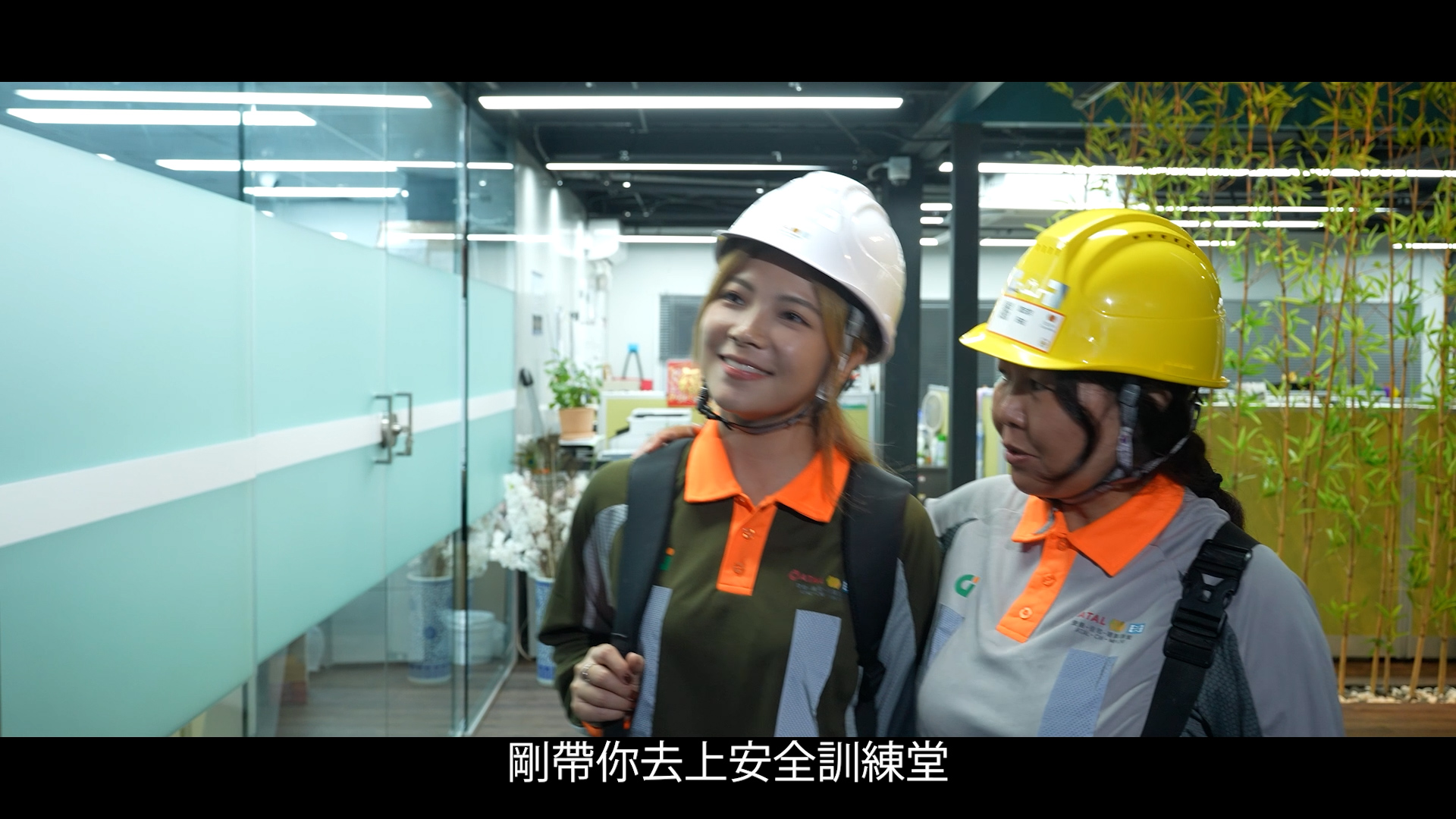 Two women wearing safety helmets and uniforms walk together inside an office building. One woman has her arm around the other. Chinese subtitles appear at the bottom of the image.