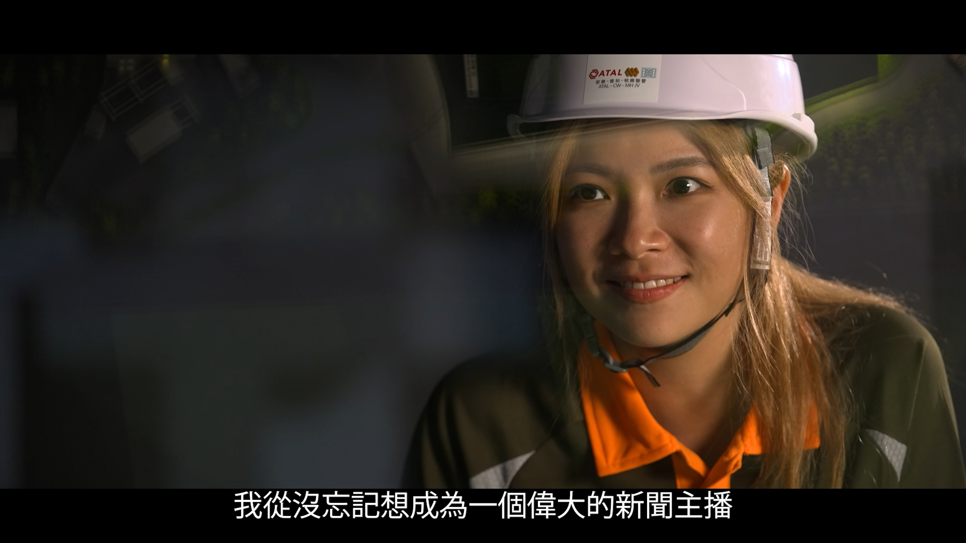 A woman wearing a white safety helmet and orange safety vest smiles while speaking. The background is dark and blurred. Chinese subtitles appear at the bottom of the image.