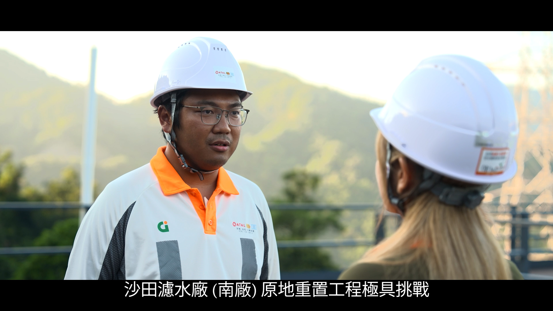 A man and a woman wearing white safety helmets and uniforms talk outdoors, with hills and a metal structure in the background. Chinese text appears at the bottom of the image.