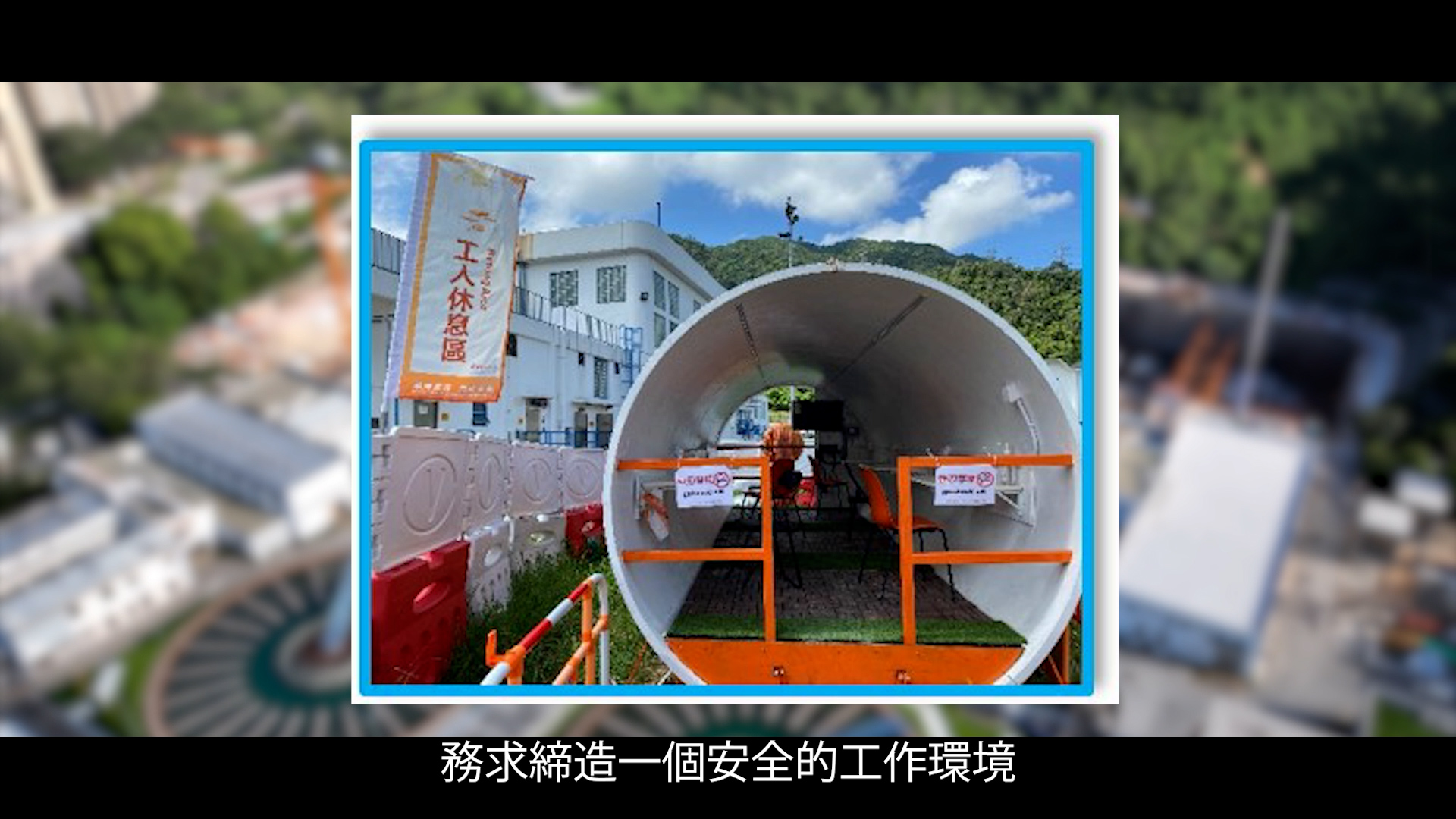 A large white tunnel segment displayed outdoors at a construction site with safety signs, scaffolding, and buildings in the background. Chinese text at the bottom reads, "務求締造一個安全的工作環境.
