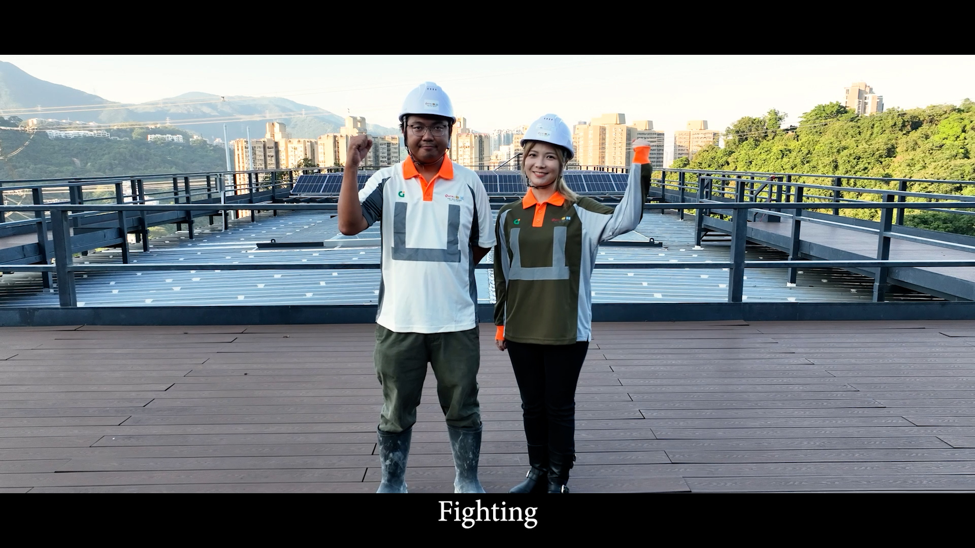 Two construction workers in hard hats raise their fists and smile, standing side by side on a rooftop with a cityscape and mountains in the background. The word “Fighting” appears at the bottom of the image.
