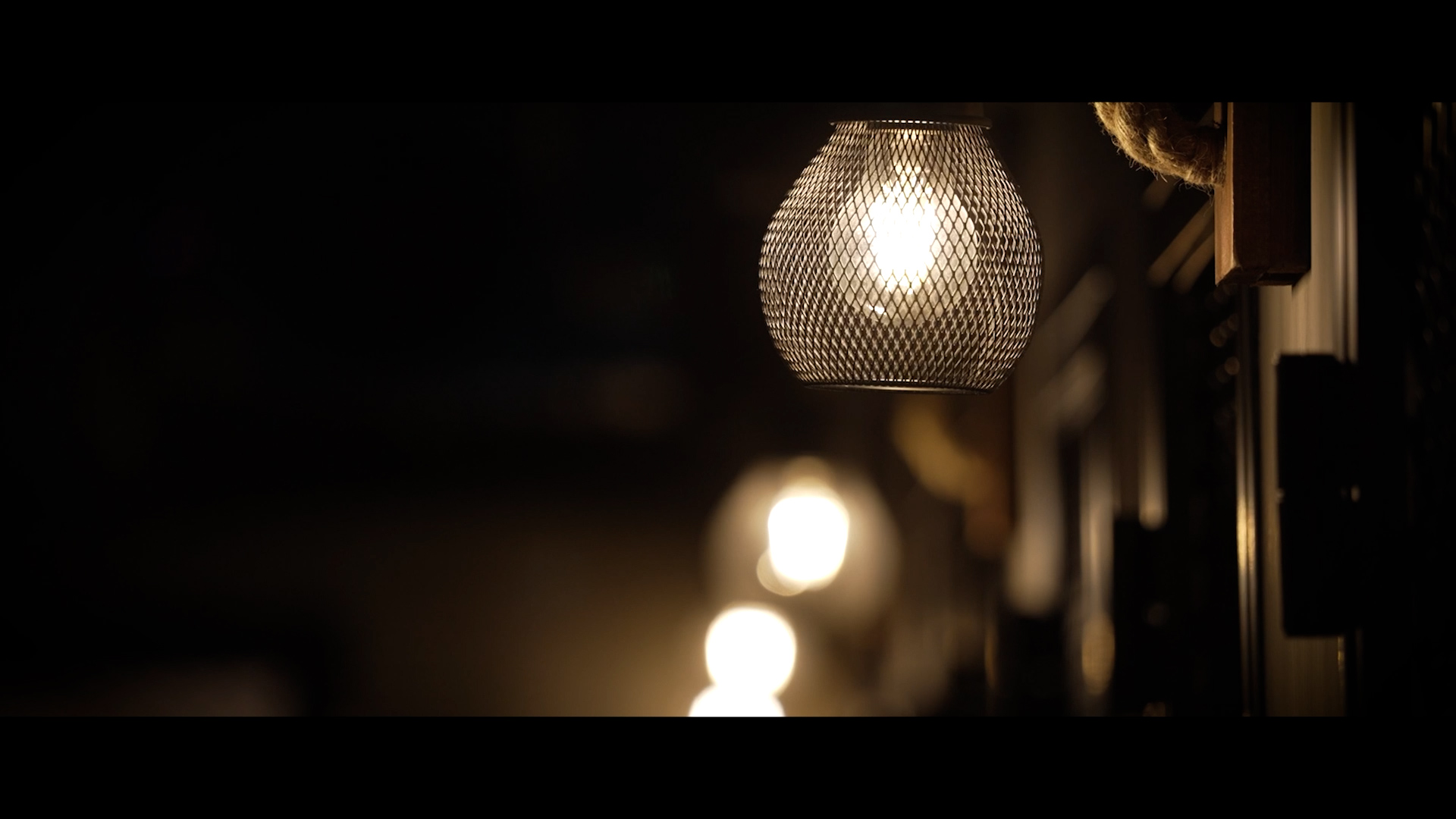 A close-up of a lit mesh lampshade hanging indoors, with two more blurred lights glowing in the background against a dark, out-of-focus setting.