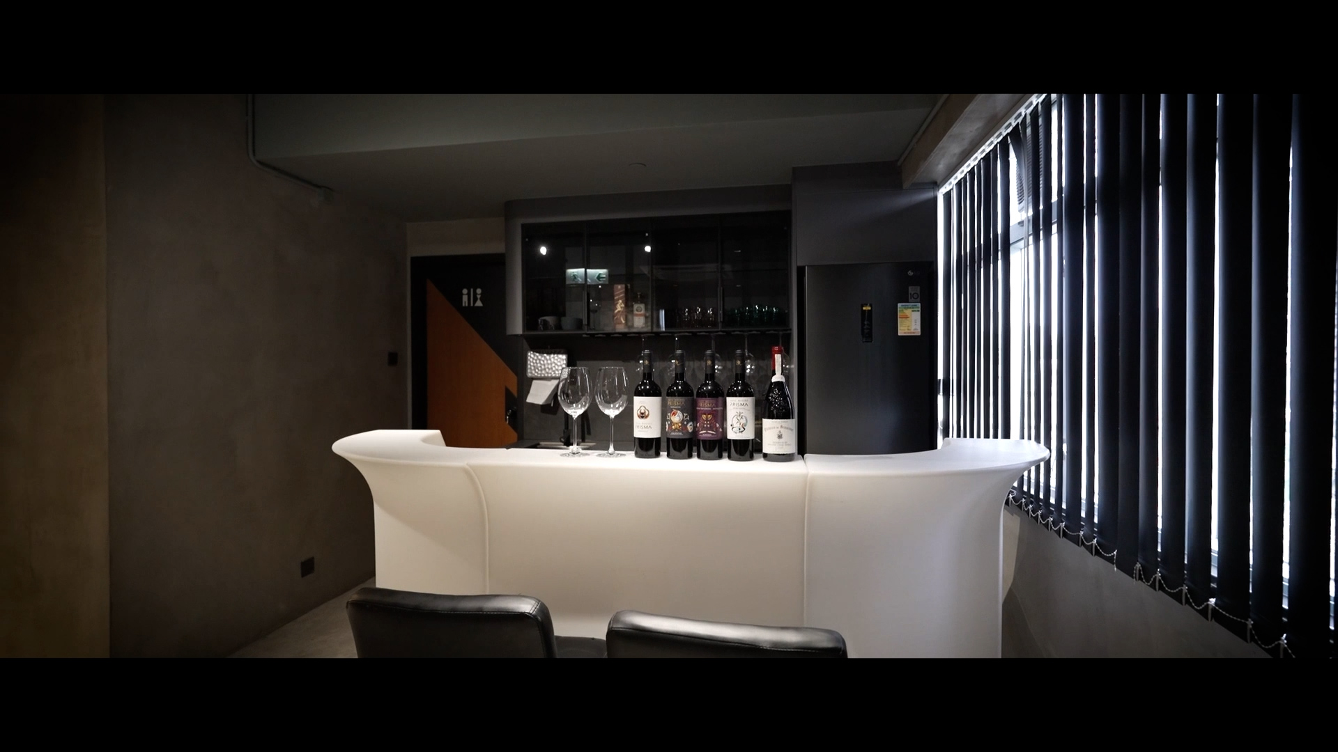 A modern home bar with a sleek white counter, several wine bottles, and glasses lined up. Behind the bar, shelves hold glassware, and a small fridge stands to the right near windows with vertical blinds. Two black chairs face the counter.
