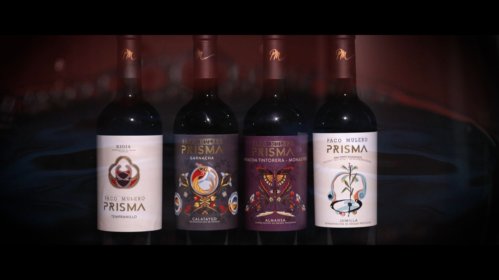 Four bottles of Paco Mulero Prisma wine stand side by side, each with a distinctively designed label in various colors and patterns, set against a dark, blurred background.