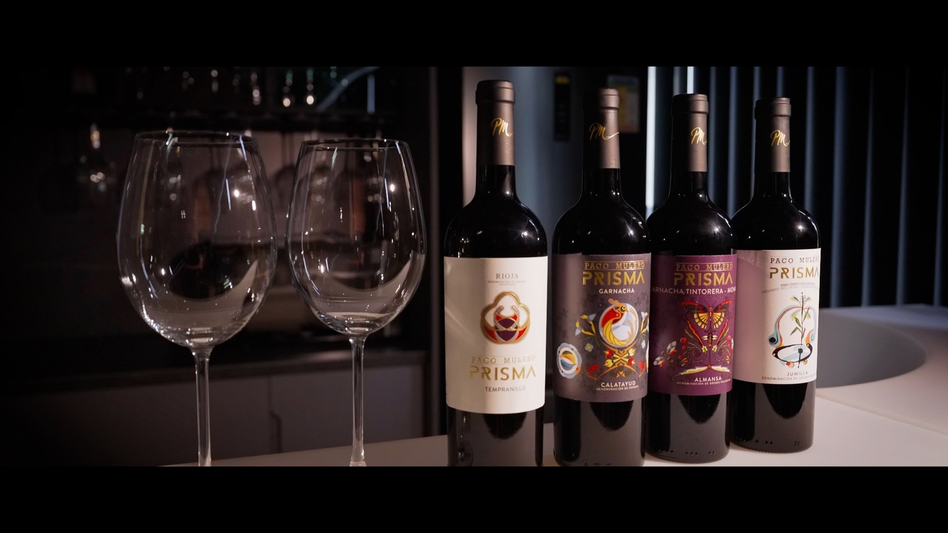 Four bottles of red wine labeled "Prisma" are lined up on a white surface beside two empty wine glasses, with a dimly lit background featuring glassware and vertical blinds.