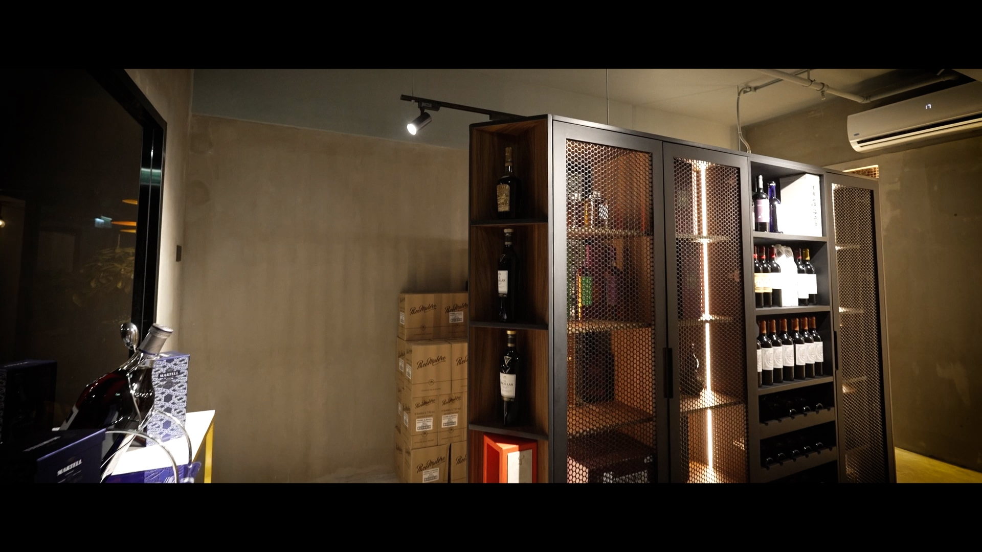 A modern wine storage room with wooden shelves filled with wine bottles, spotlights above, some stacked cardboard boxes on the left, and an air conditioner mounted on the wall. The walls are concrete and minimally decorated.
