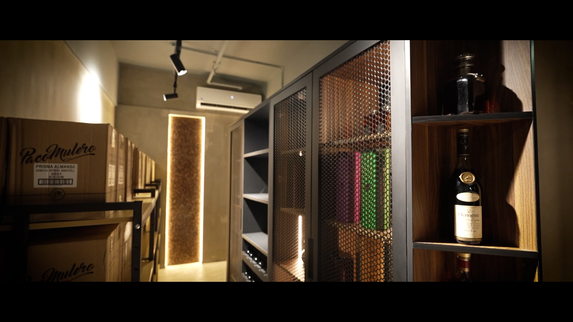 A modern wine storage room with wooden shelves, metal mesh doors, cardboard boxes, and bottles of wine, softly lit with warm lighting and an air conditioner mounted on the back wall.