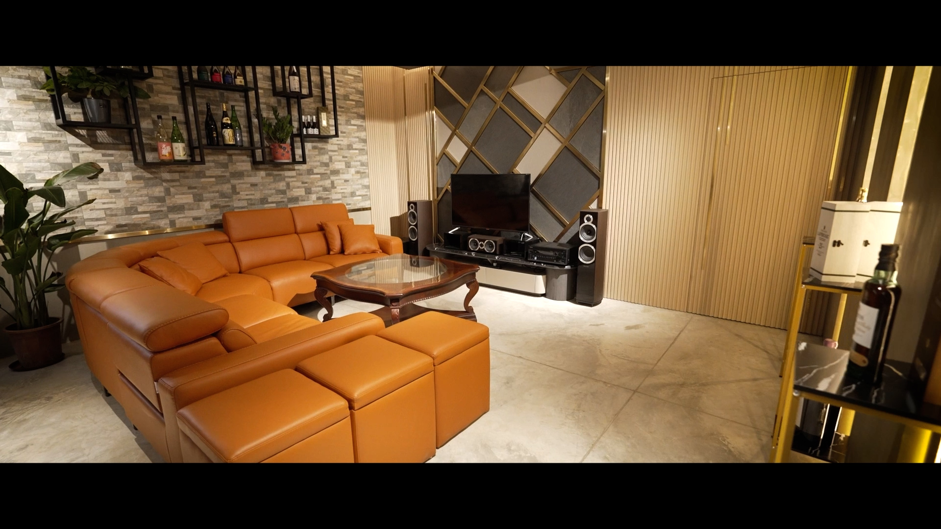 A modern living room with an orange sectional sofa, glass coffee table, TV with speakers, decorative shelves with bottles, wall plants, and patterned accent walls. The space appears cozy and stylish.