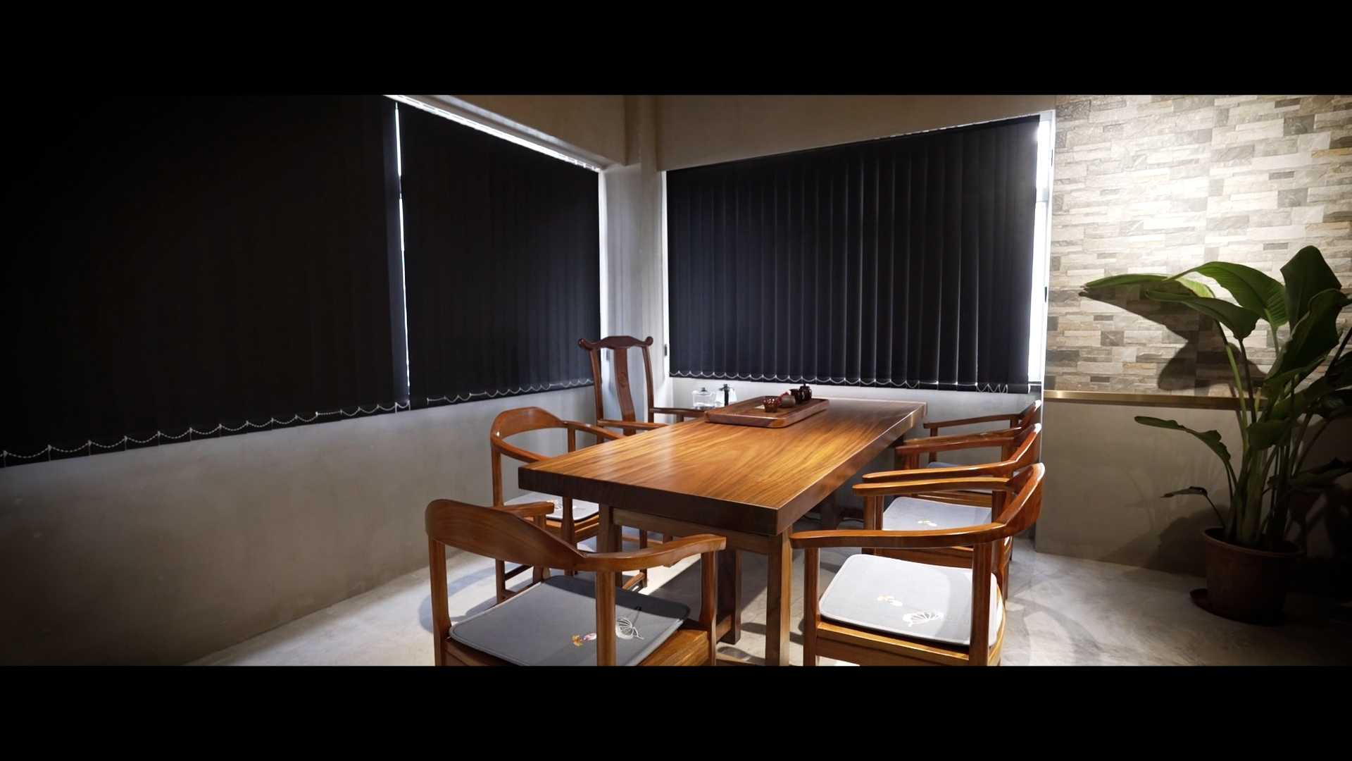 A modern dining area with a wooden table and five wooden chairs, set near two windows with closed black blinds. A potted plant stands by a textured wall in the corner. The room has a minimalist, cozy feel.