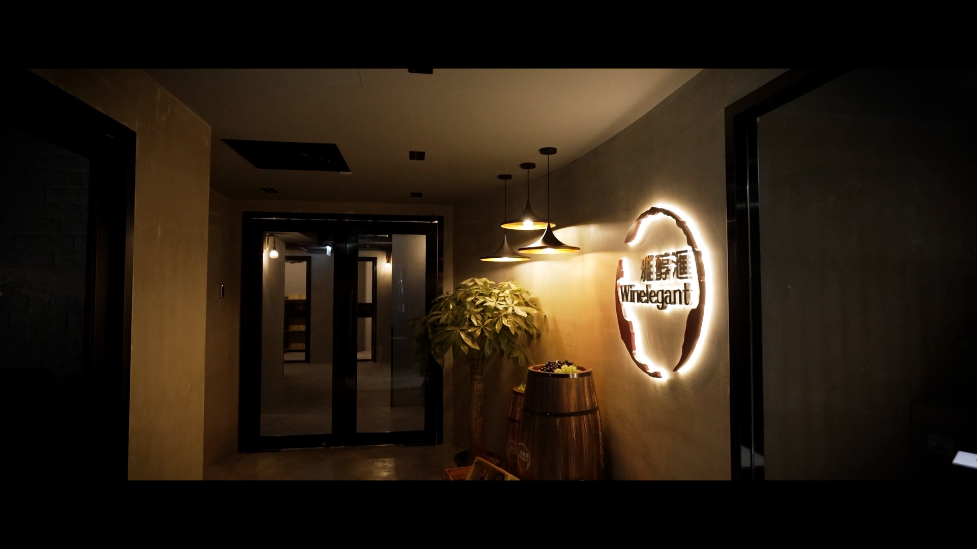 A dimly lit hallway with a glowing wall sign reading "Wine&gant" in English and Chinese characters, three hanging pendant lights, a potted plant, and a wooden barrel near a glass door entrance.