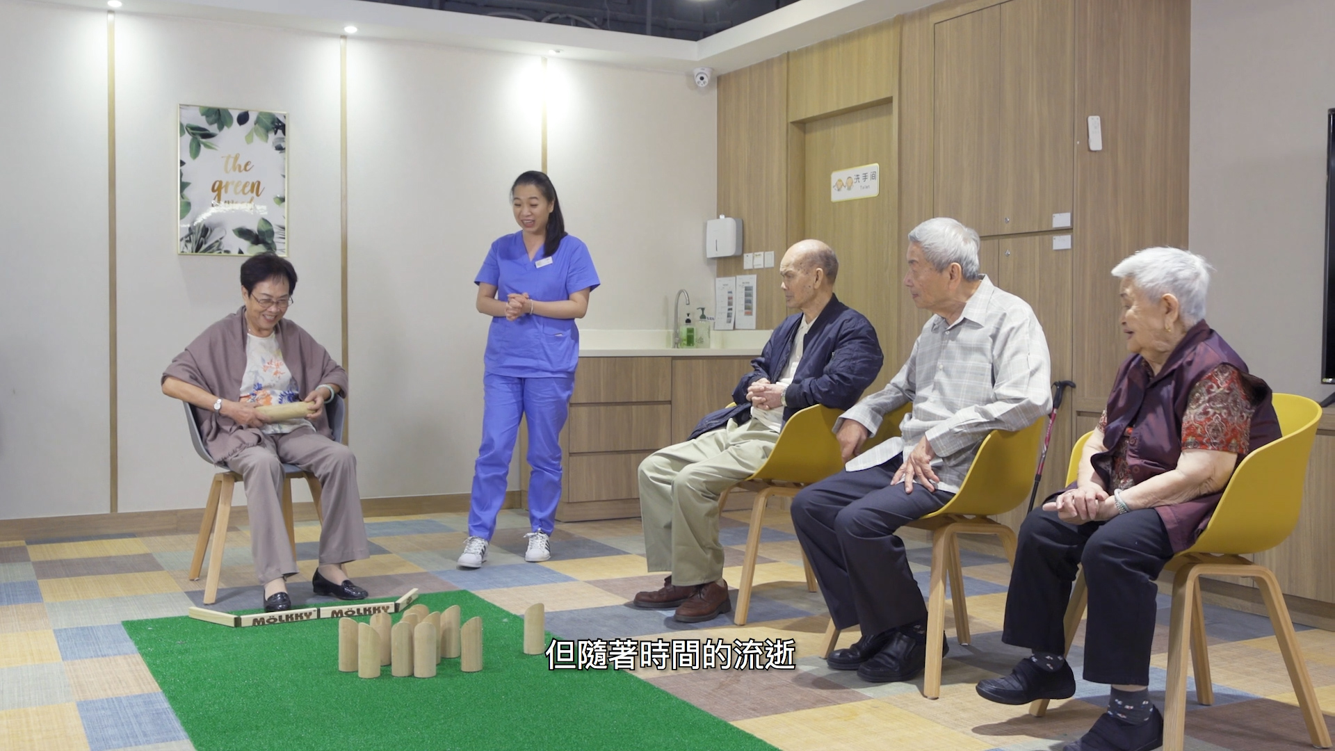 A group of elderly people sit in chairs, watching a woman play a domino game on a green mat, with a staff member standing nearby in a room labeled "The Green." Subtitles in Chinese are visible at the bottom.