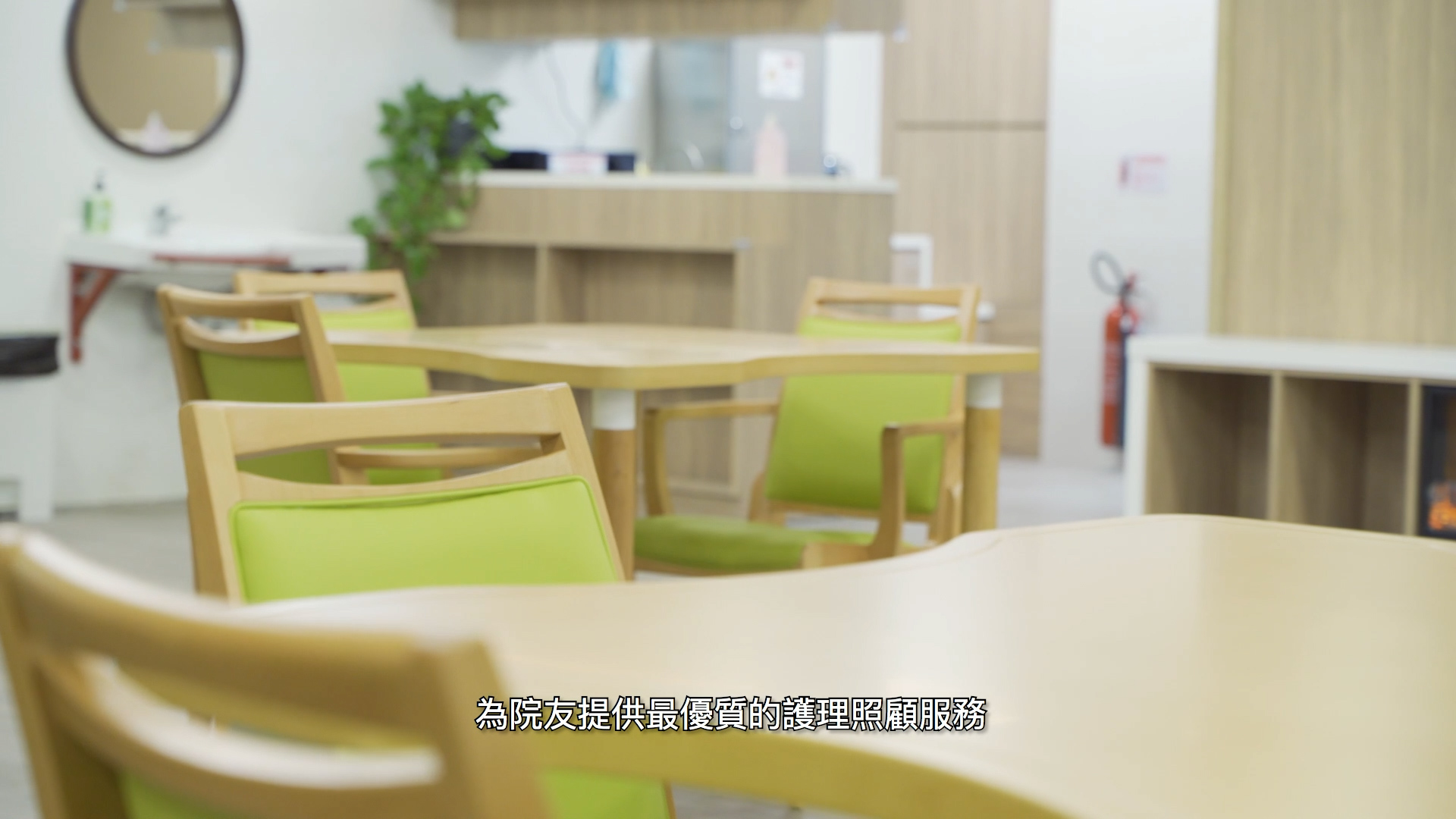 A modern, bright café or dining area with light wooden tables and green chairs. In the background are shelves, a mirror, and a fire extinguisher. Chinese text appears at the bottom center of the image.