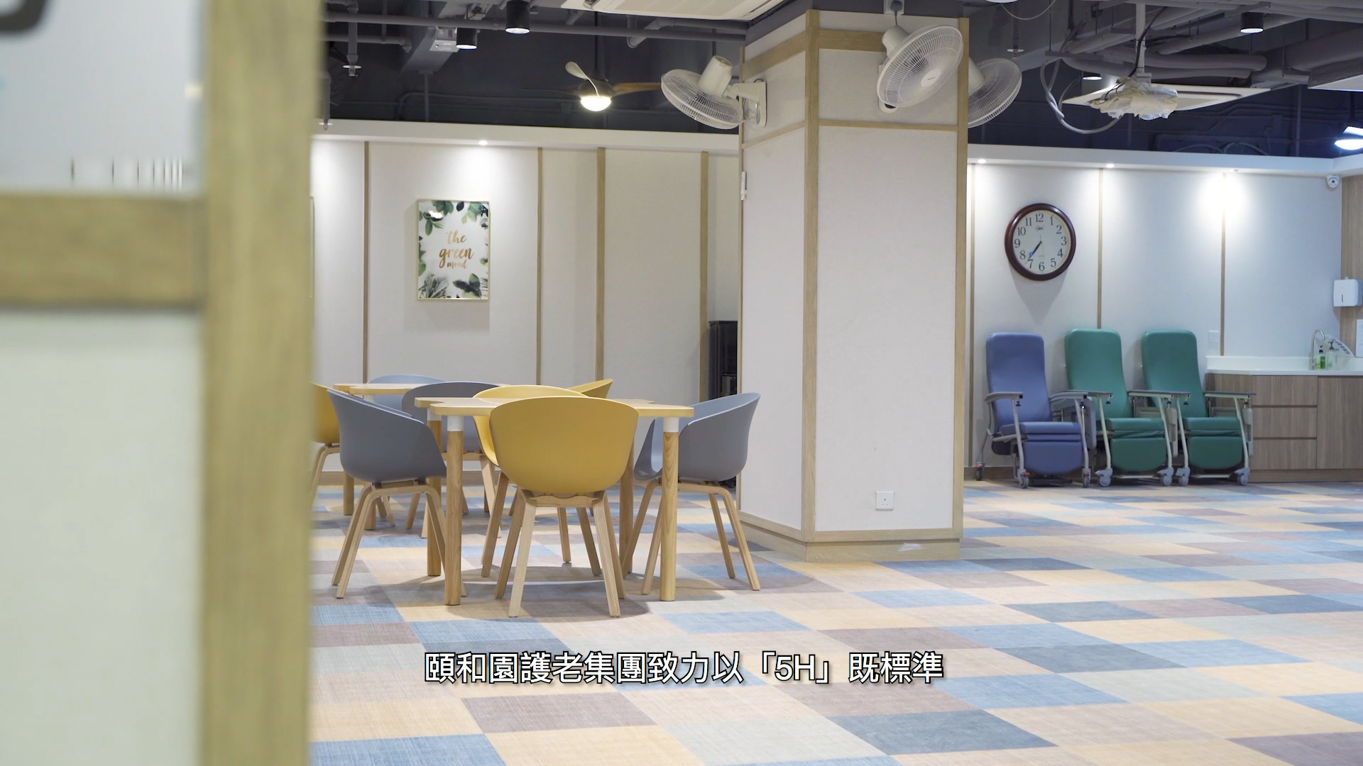 A modern, spacious room with colorful chairs around a table, several recliner chairs along the wall, a large clock, and soft lighting. Chinese text is displayed at the bottom of the image.