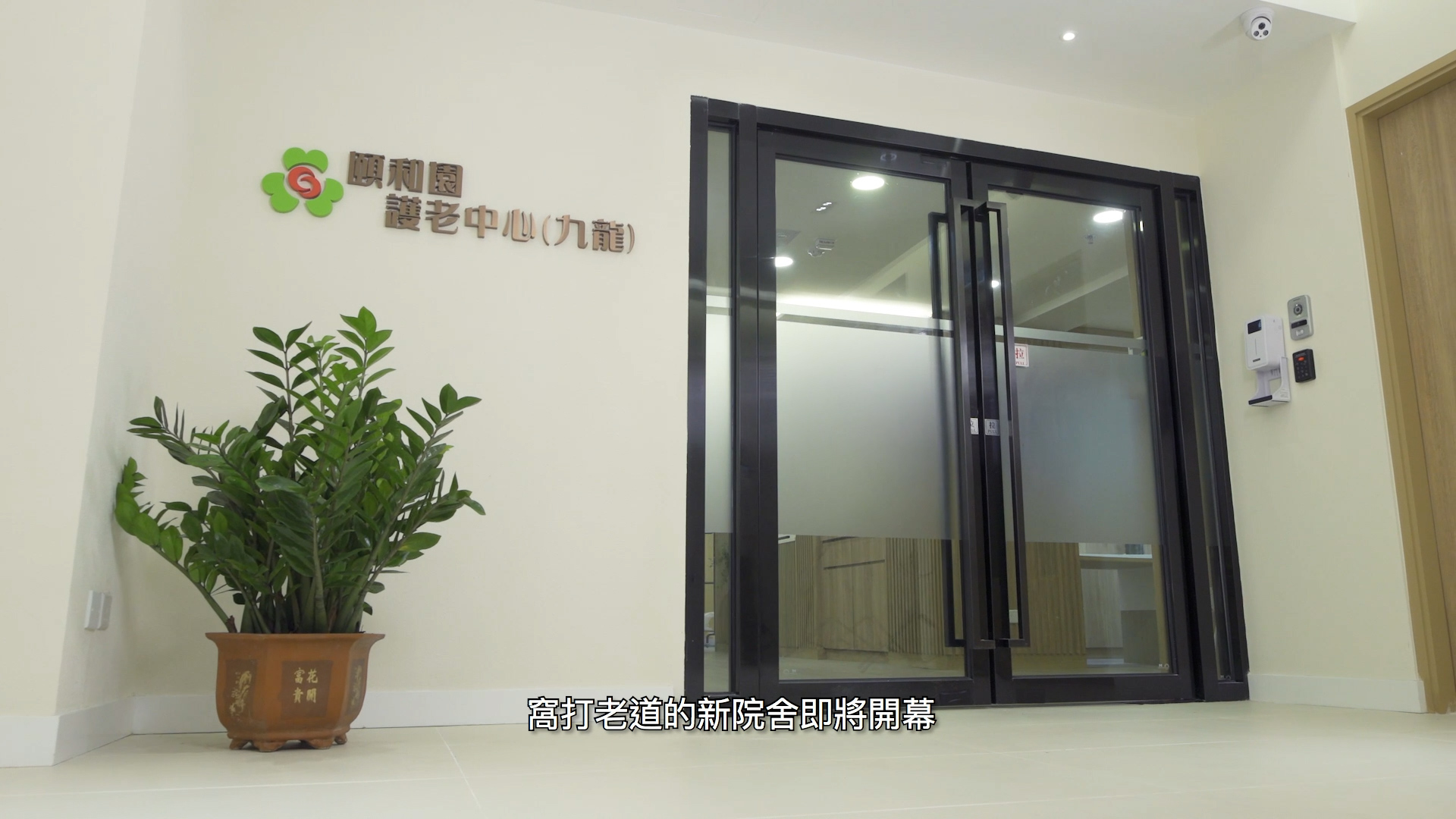 A modern glass door entrance to an elderly care center, with a potted plant on the left and the center’s name in Chinese on the wall. Chinese subtitles appear at the bottom of the image.