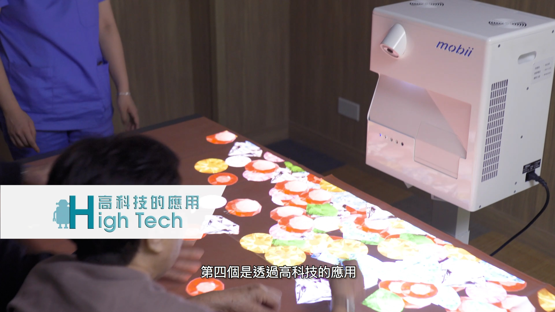 An elderly person interacts with a colorful projected display on a table while a caregiver stands nearby. A machine labeled "mobii" projects images. Chinese text and "High Tech" are overlaid in the bottom left corner.