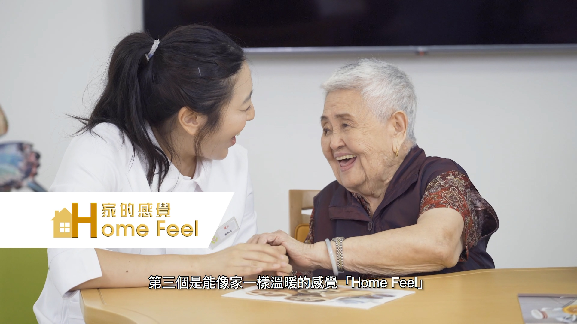 A young woman and an elderly woman sit at a table, smiling and holding hands warmly. The elderly woman looks joyful. The image includes the "Home Feel" logo and Chinese text at the bottom.
