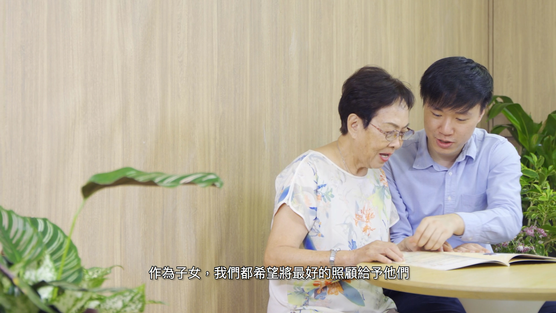 A young man sits beside an elderly woman, helping her read a book at a wooden table in a bright room with green plants; Chinese text appears at the bottom of the image.