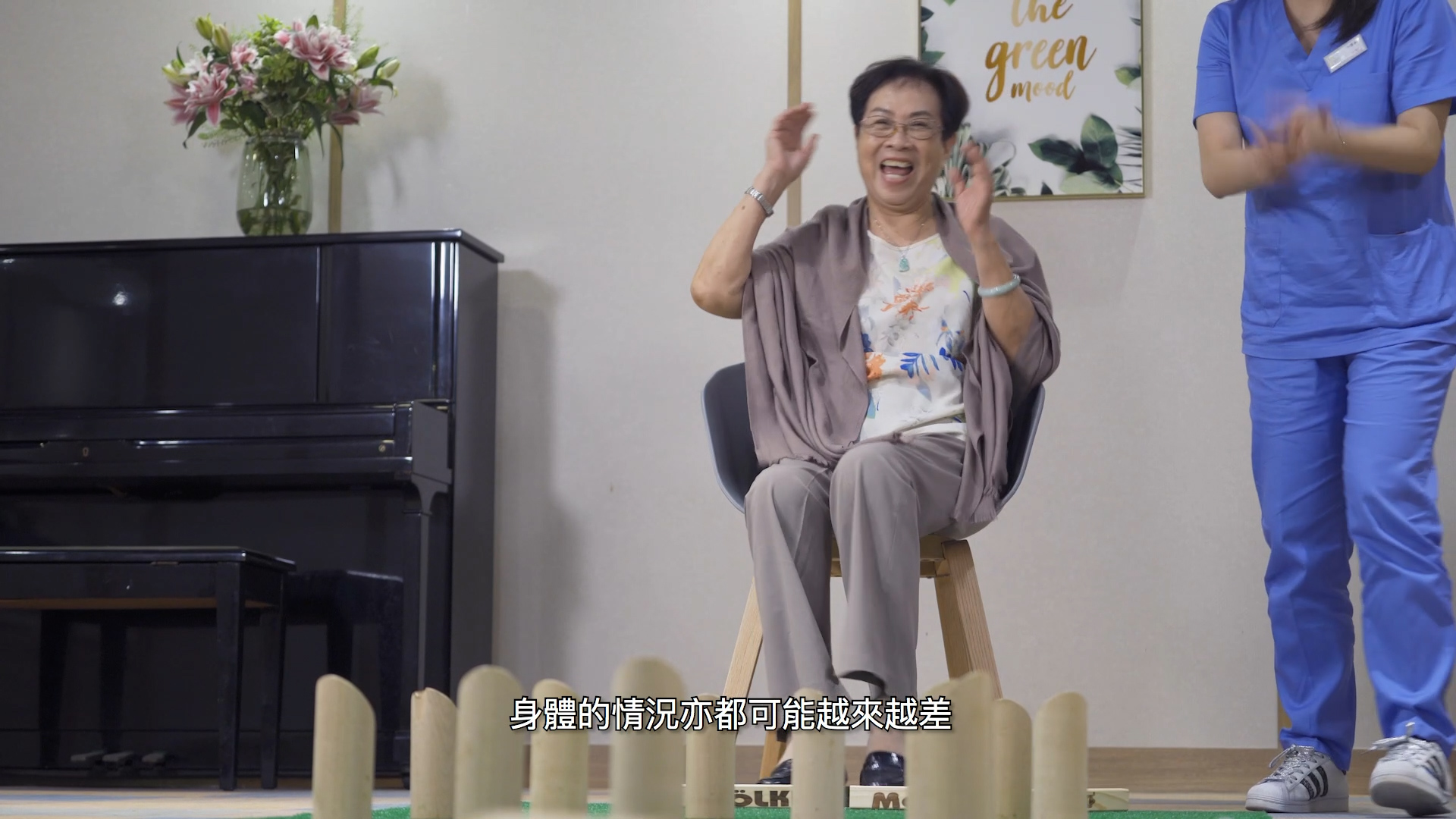 An elderly woman sits on a chair, smiling and clapping her hands, next to a person in blue scrubs. Wooden pins are arranged in the foreground, and a piano and flowers are seen in the background. Chinese subtitles appear at the bottom.