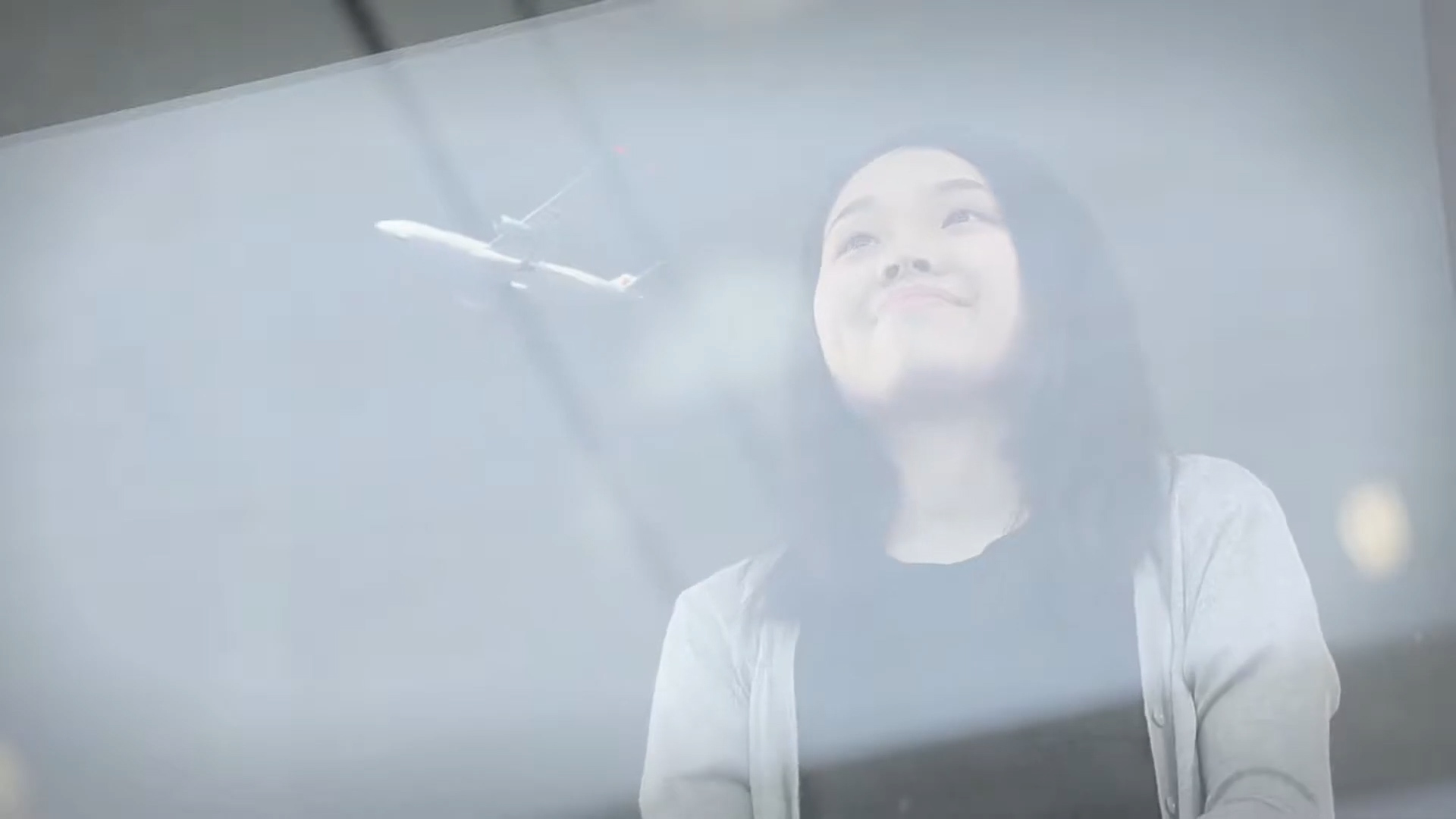 A woman smiles softly while looking up, with the faint image of an airplane flying in the sky above her, seen through a glass window.