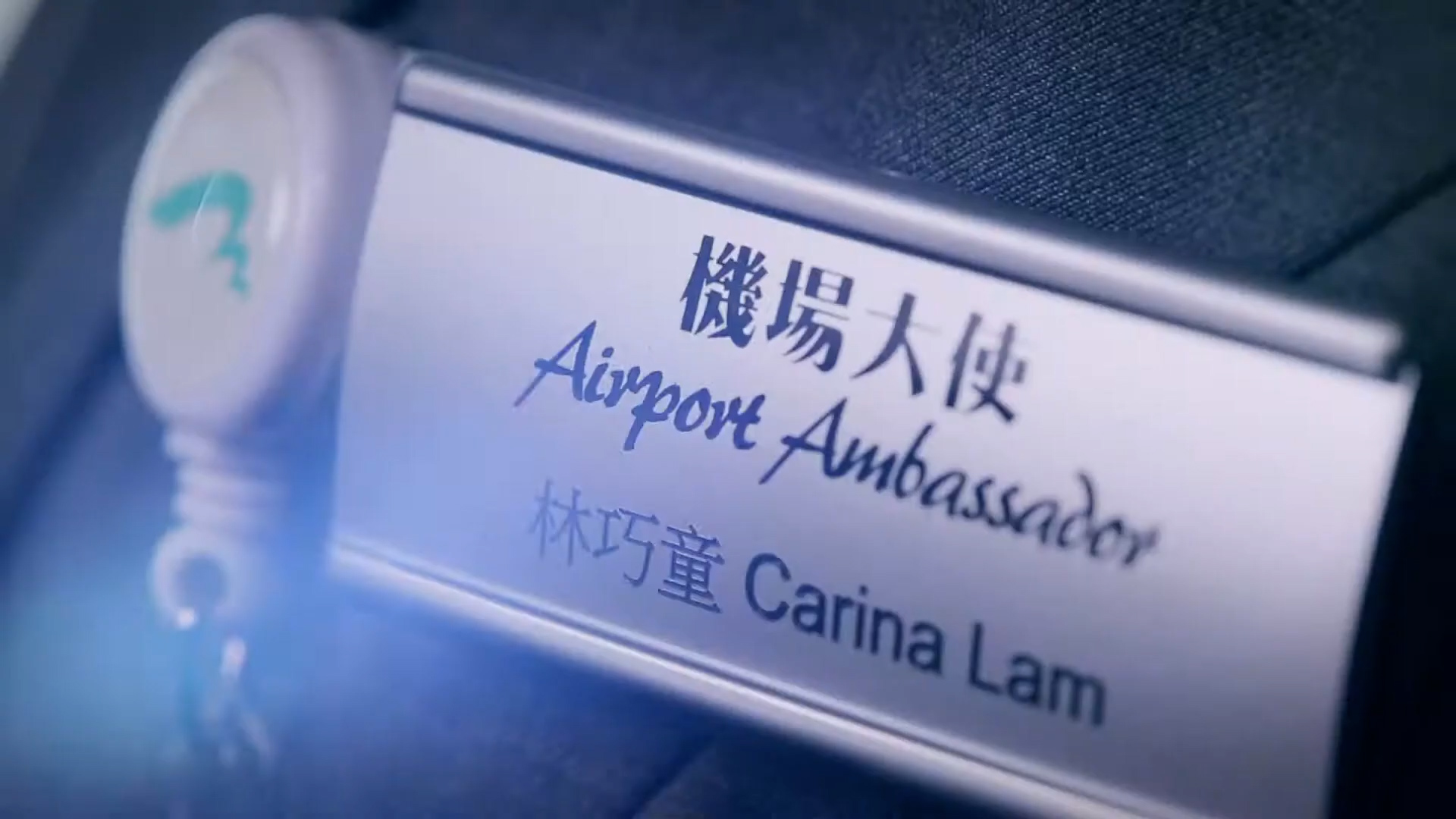 Close-up of a name badge reading "Airport Ambassador" with the name "Carina Lam" and additional text in Chinese, pinned to a dark-colored uniform.