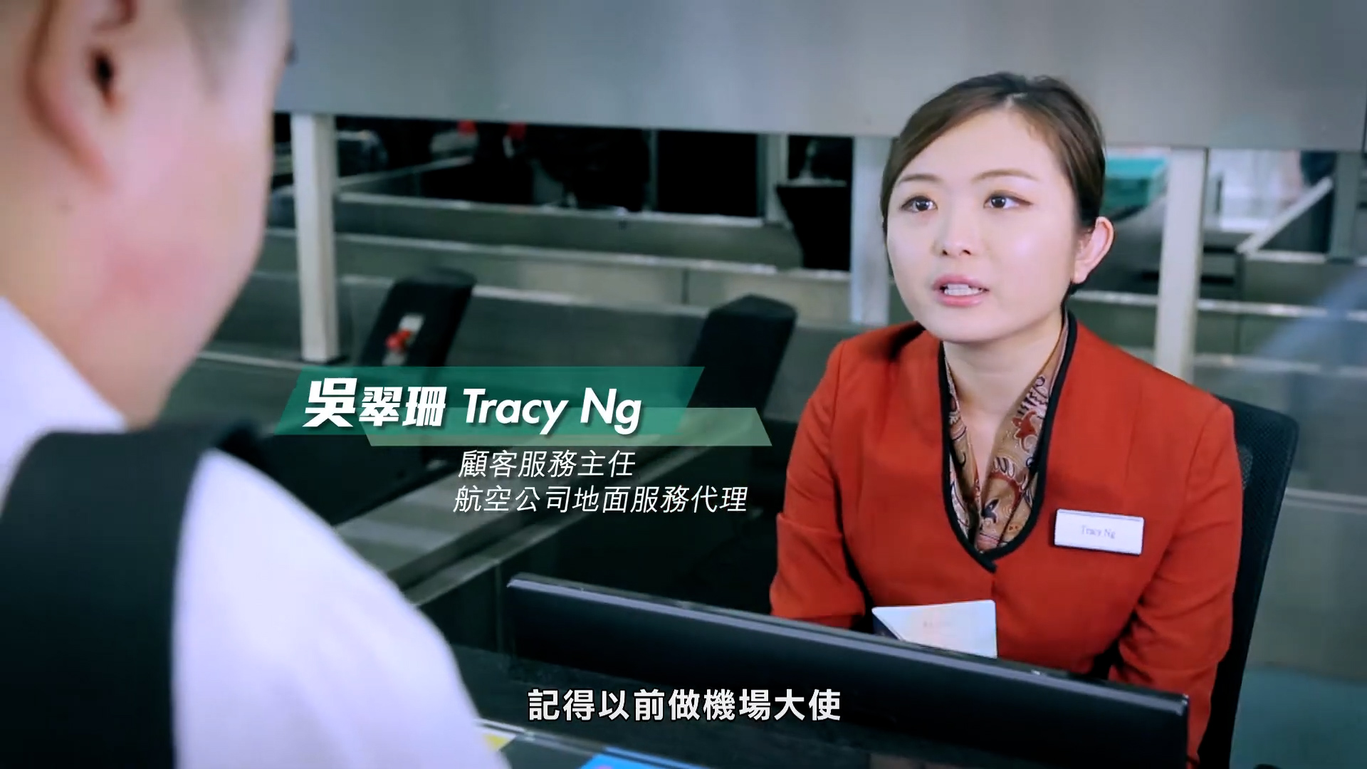 A woman in a red blazer with a name tag speaks to a customer at an airline service counter. Text on screen identifies her as Tracy Ng, with additional information in Chinese, and Chinese subtitles appear at the bottom.