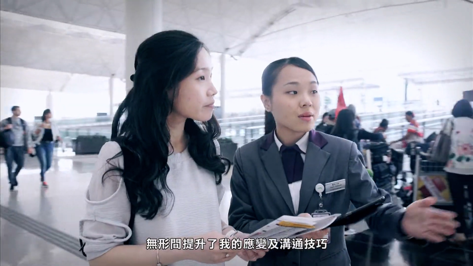 Two women stand indoors; one is dressed professionally and holding a clipboard, appearing to assist or guide the other woman. Background shows a modern, spacious building. Chinese subtitles are present at the bottom of the image.