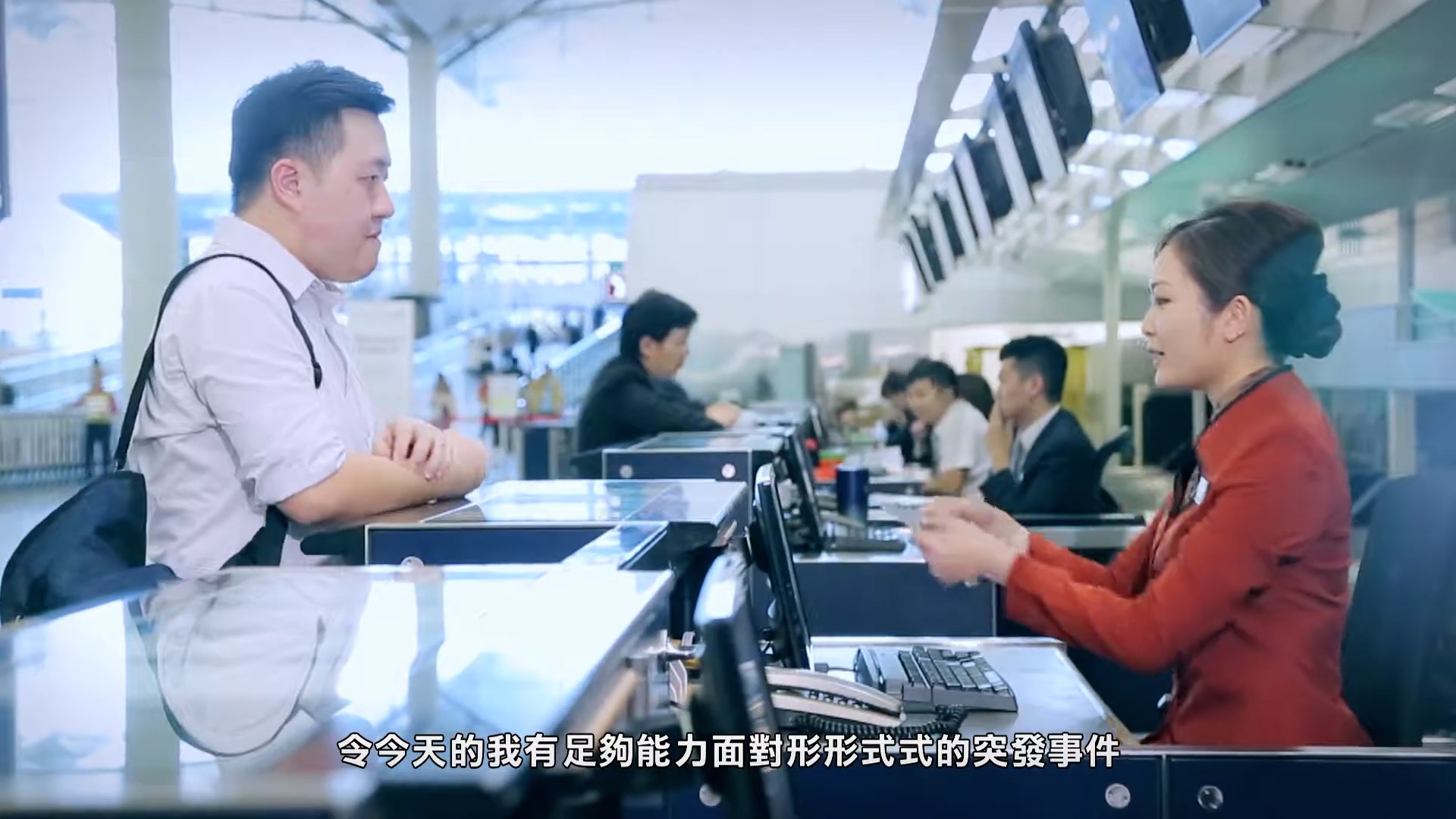 A man stands at an airport check-in counter speaking with a female staff member in uniform. Other agents and passengers are visible in the background. Chinese subtitles are displayed at the bottom of the image.