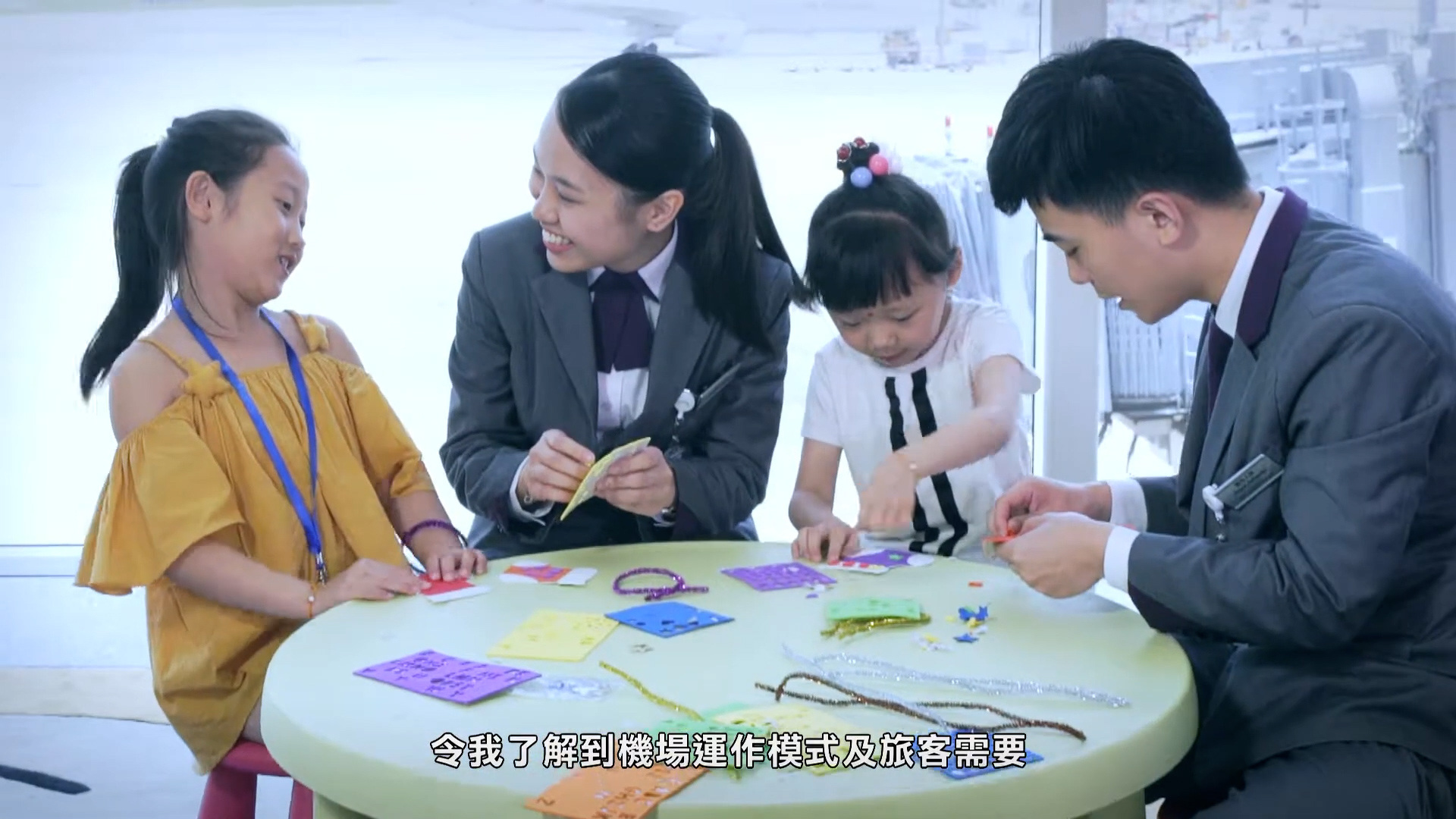 Two adults in suits and two young girls sit around a round table, smiling and making crafts with colorful paper and beads. Chinese subtitles appear at the bottom of the image.