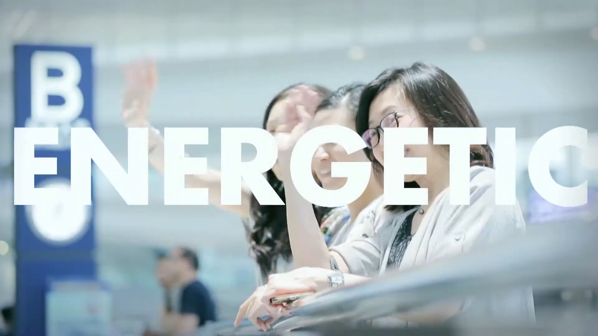 Three people stand indoors near a railing, waving enthusiastically. The word "ENERGETIC" in large white letters is superimposed over them. The background is blurred, showing a blue sign with a white letter "B.