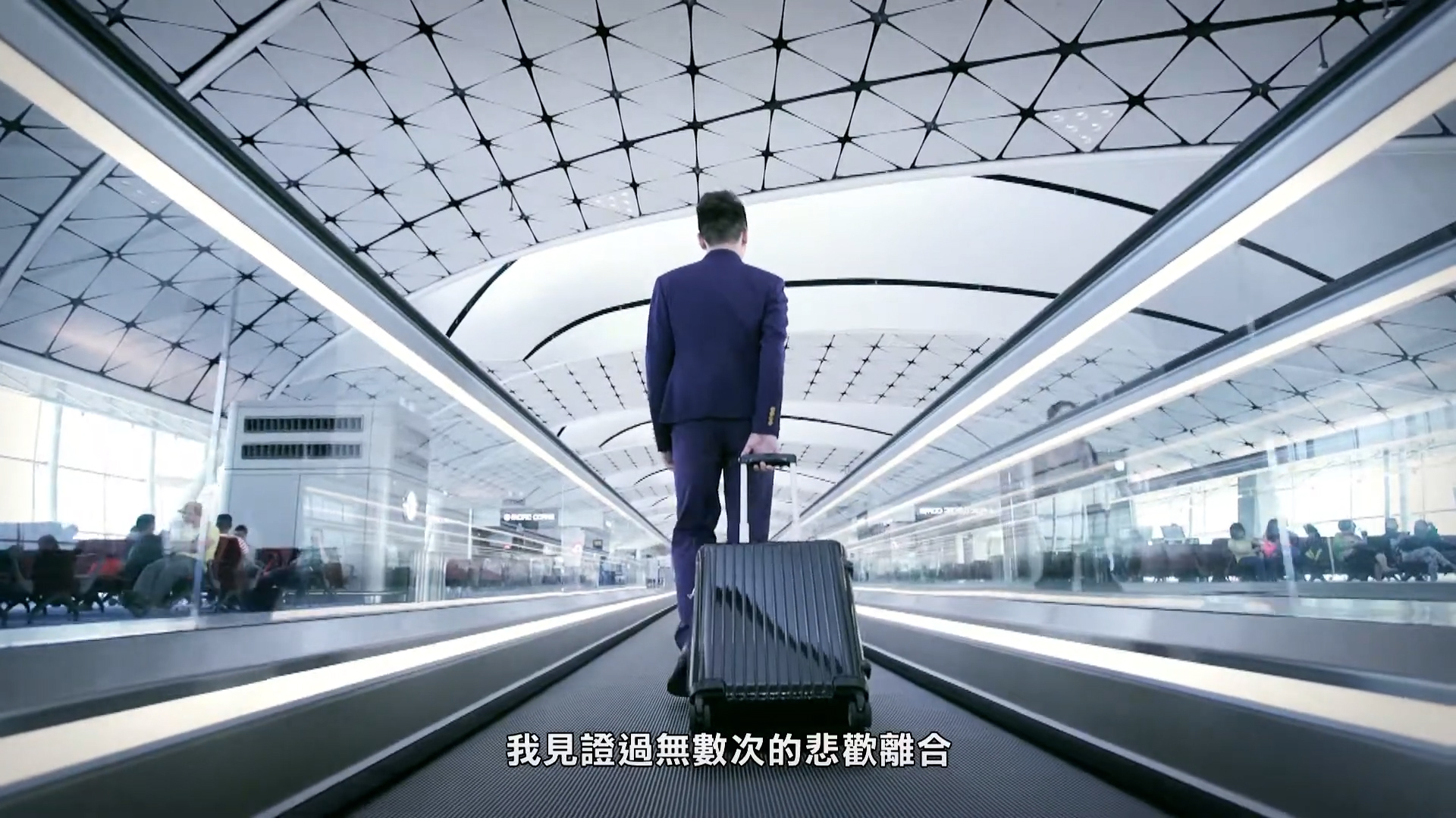 A man in a blue suit walks with a rolling suitcase on a moving walkway in a modern airport, with Chinese subtitles at the bottom of the image.