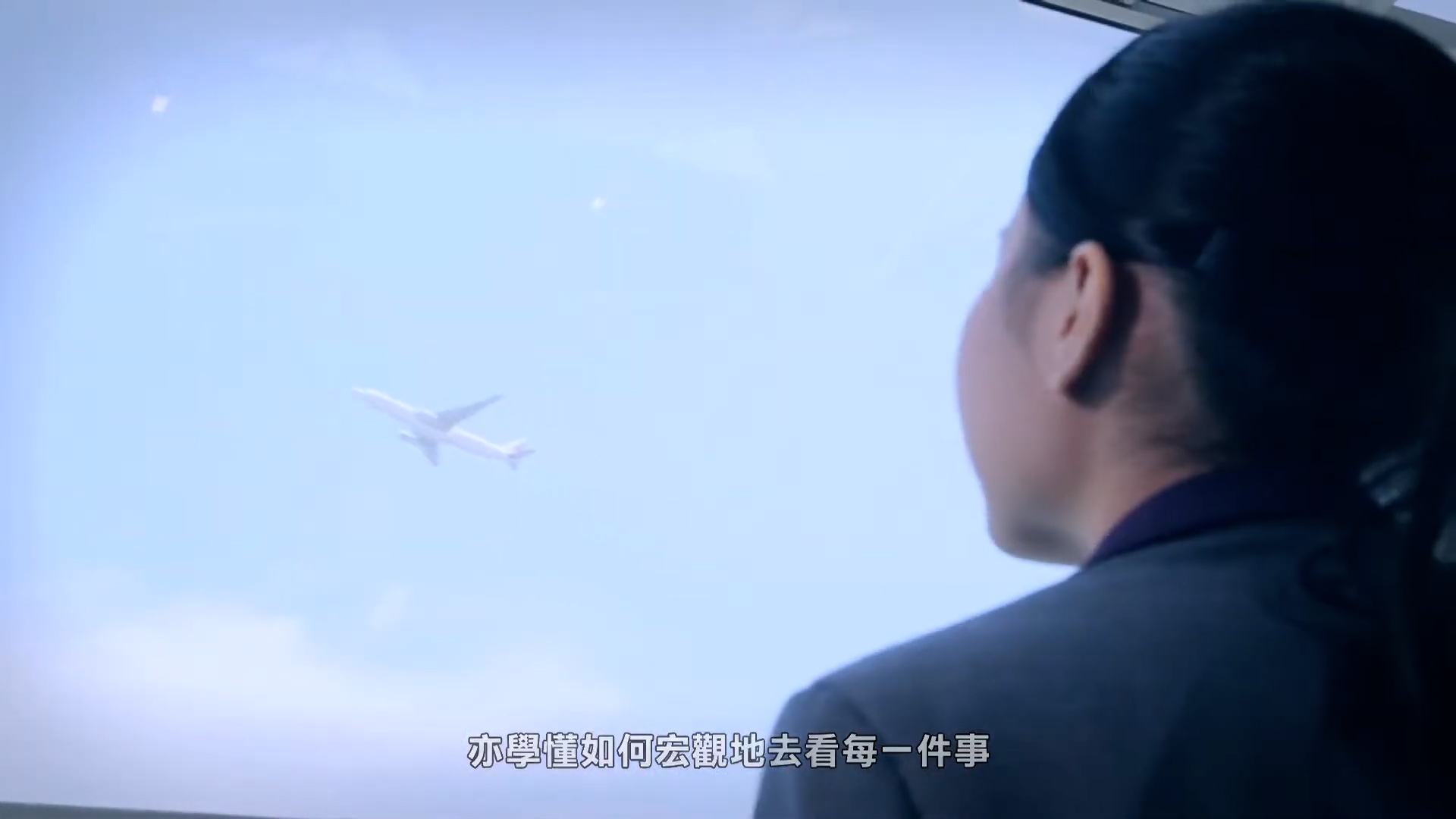 A woman with dark hair, wearing a suit, looks out a window at an airplane flying in a blue sky. Chinese subtitles are visible at the bottom of the image.