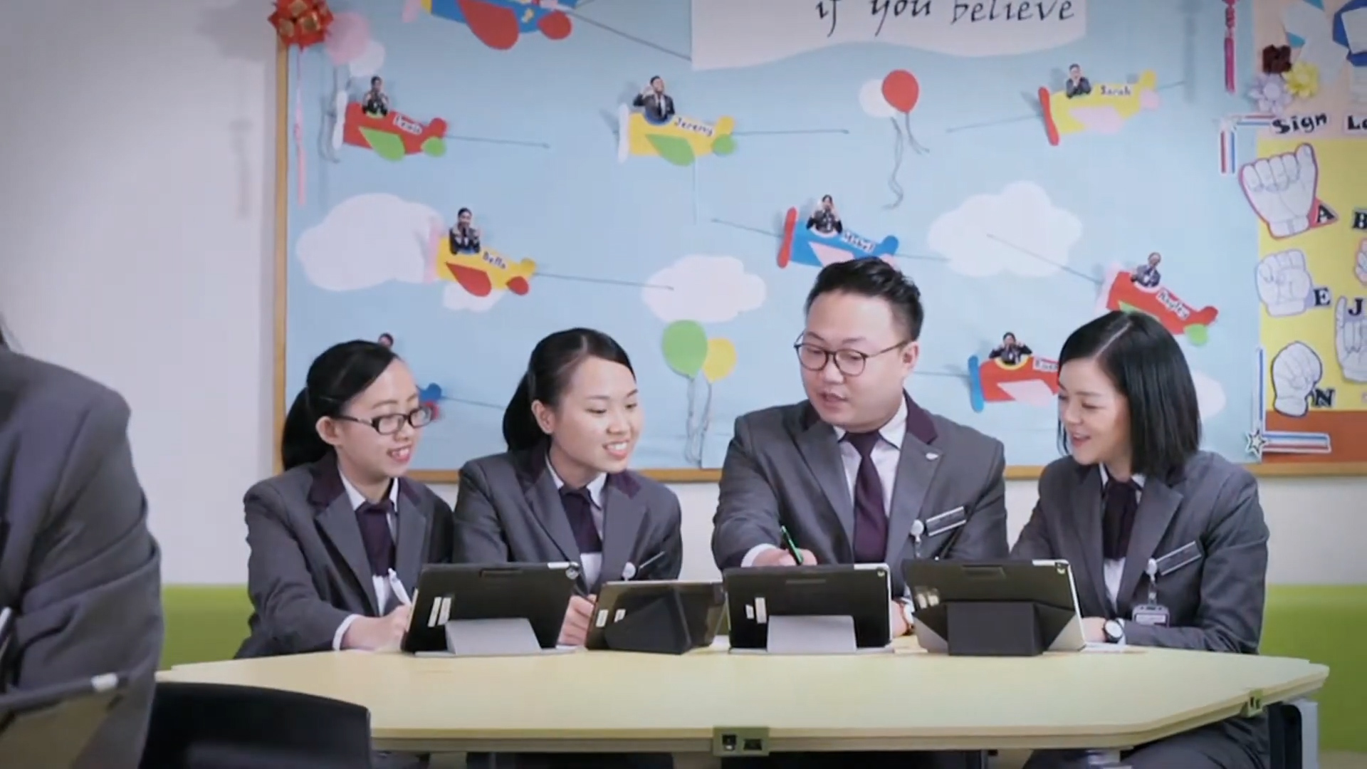 Four people in gray uniforms sit at a round table, using tablets. They are smiling and talking. Behind them is a colorful wall decorated with paper airplanes and photos of people, creating a lively atmosphere.